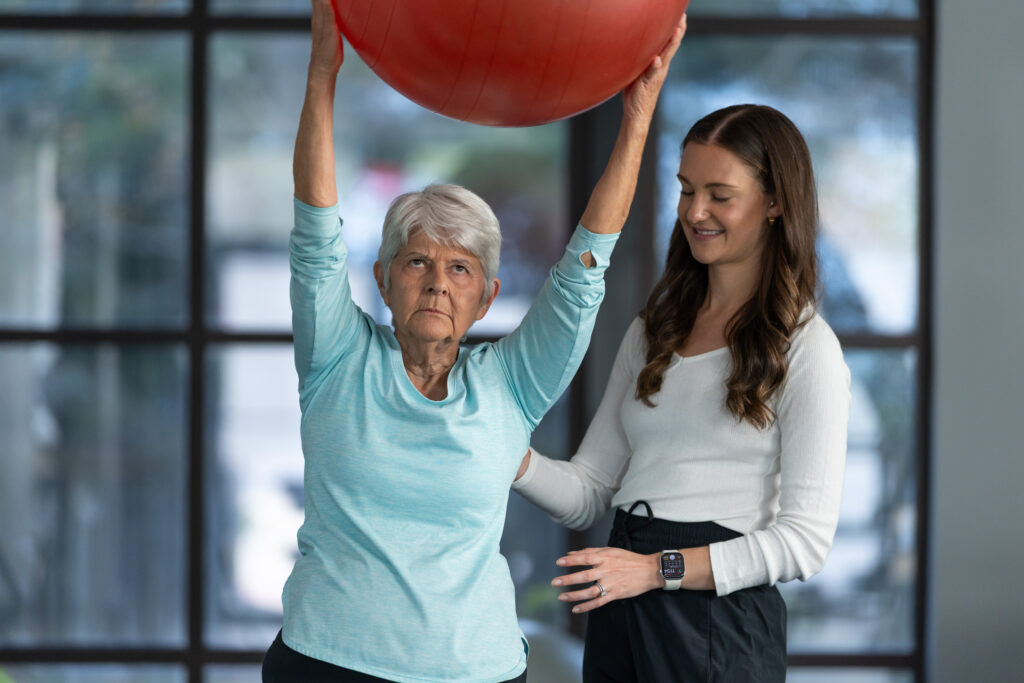 Woman holding exercise ball while female physical therapist supervises