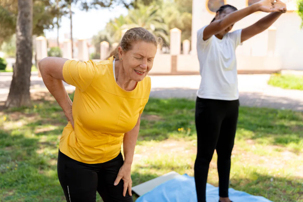 An older woman in a yellow shirt holds her lower back in pain, eyes closed, while a younger woman stretches in the blurred background outdoors.