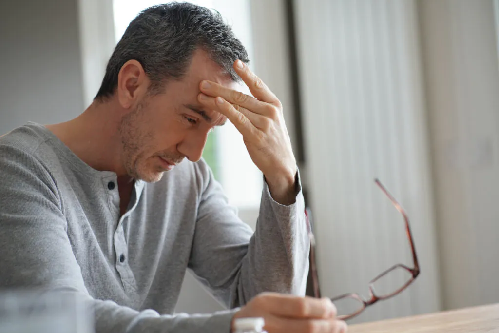 A man with graying hair holds his forehead with one hand and glasses with the other, looking down in distress.