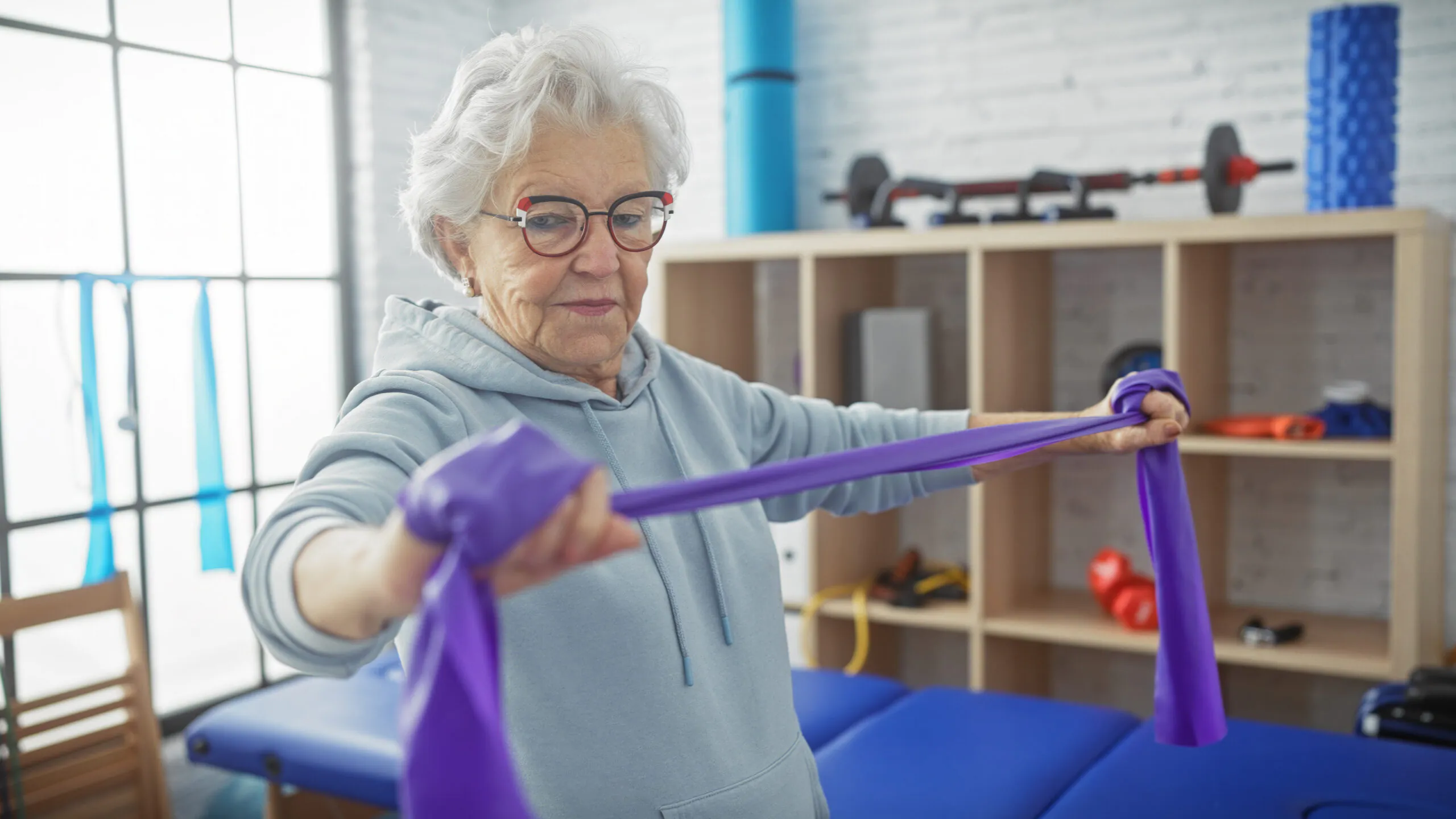 An elderly woman with white hair and glasses stretches a purple resistance band horizontally in a gym setting.