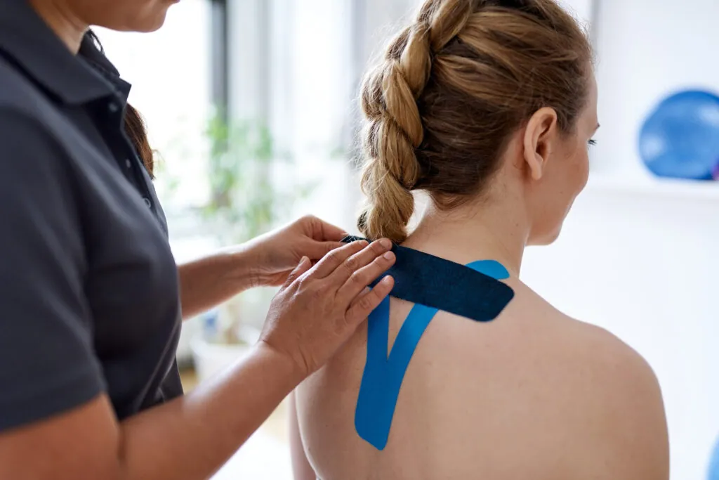 Candid image of a patient during appointment with a professional physiotherapist in design kinesiotherapy clinic stretching medical kinesio tape for neck and shoulder pain.