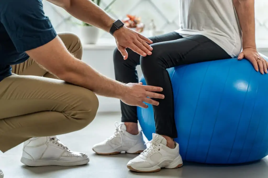 Close-up of physiotherapist supporting senior woman in rehabilitation leg exercise on balance ball during physical therapy session.