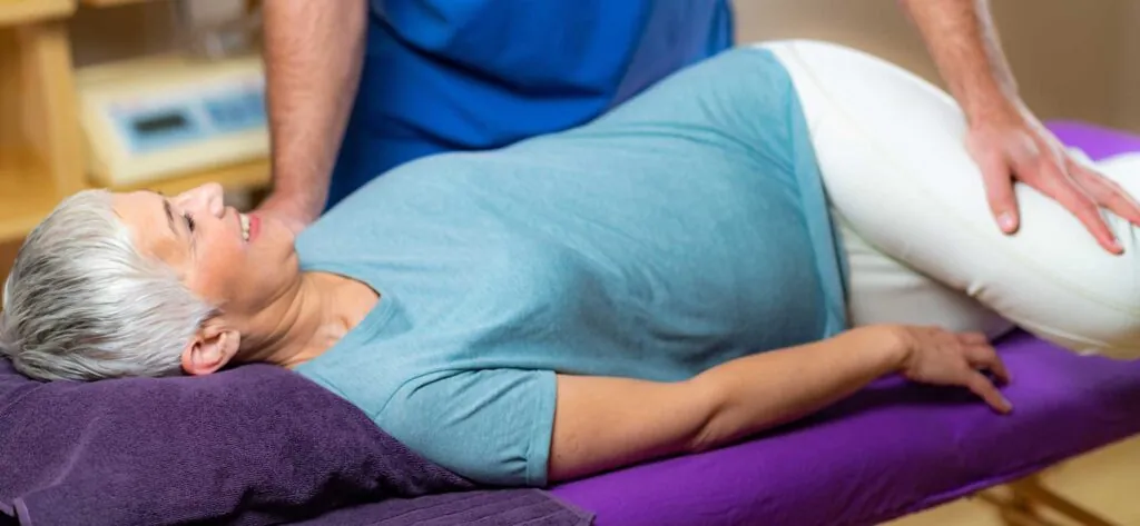 An older female patient stretching on an exercise table with a physical therapist helping her