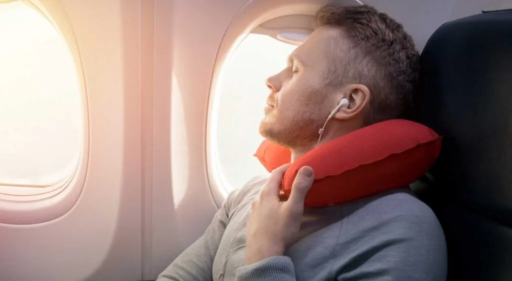 a plane passenger with a red neck pillow, sleeping next to the plane window