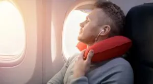 a plane passenger with a red neck pillow, sleeping next to the plane window