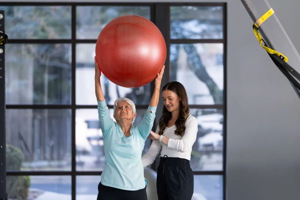 an elderly patient lifting a ball with a physical therapist adjusting their backs position
