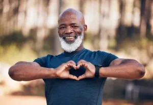 Black man, fitness and heart portrait for exercise, fitness and health while outdoor with a smile. Healthy senior person in nature forest happy with hand emoji sign for workout and cardio training.