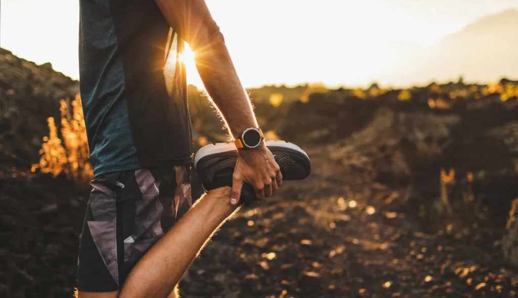 Male runner stretching leg and feet and preparing for running outdoors. Smartwatches or fitness tracker on hand. Beautiful sun light on background. Active and healthy lifestyle concept.