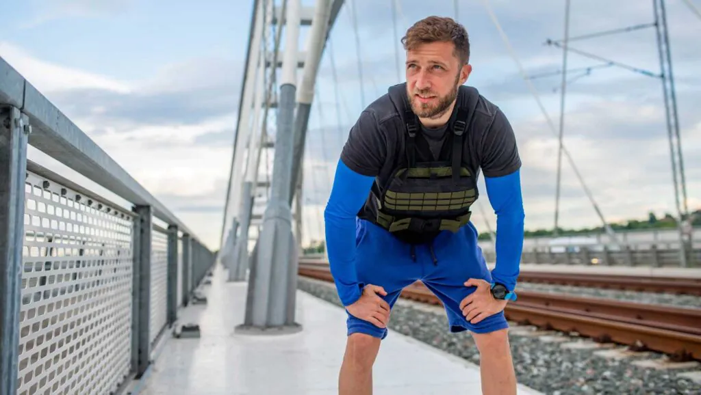 a young runner wearing a weighted vest in resting position with his hands resting on his knees. 