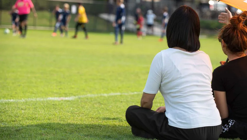 Back view of a supportive Asian mother watching her son playing soccer at a local school tournament in the afternoon.