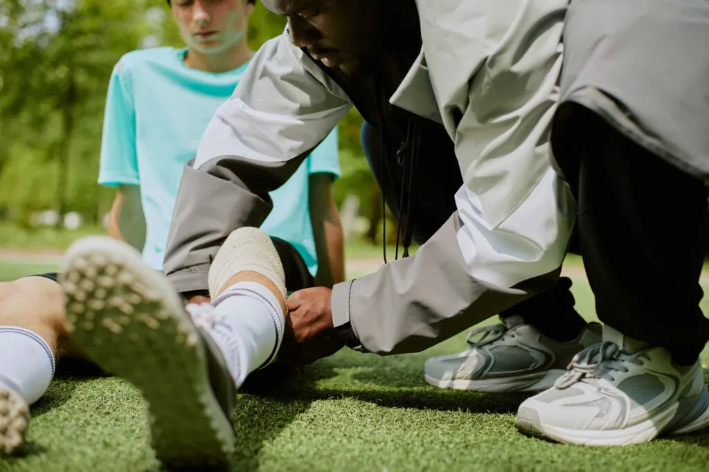 Black man assisting injured teenager on sports field, kneeling and examining leg while another teenager sits nearby observing situation, outdoor setting with green grass visible