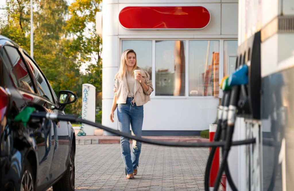 a woman holding a coffee while filling up her car at a gas station