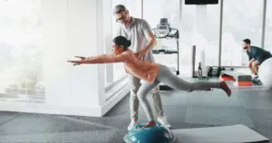 Physiotherapy, woman and personal trainer in gym for balance, rehabilitation and stability training. Man, client and half ball in fitness center for core strength, yoga practice and help in exercise.