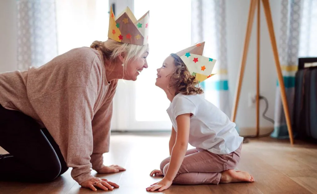 A portrait of small girl and grandmother with paper crown having fun at home.