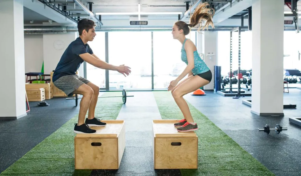 Beautiful young fit couple exercising in modern crossfit gym, doing box jumps.