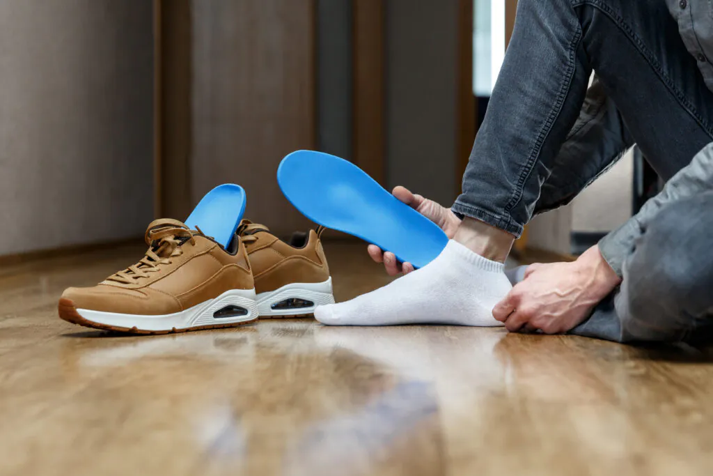 A person inserting a bright blue orthopedic insole into a brown sneaker on a wooden floor. Another brown sneaker with an insole is beside it.
