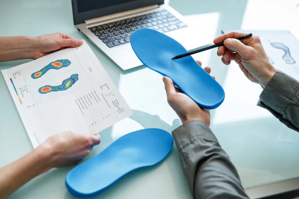 Two people examining foot pressure maps and blue custom shoe insoles on a table, with a laptop in the background.
