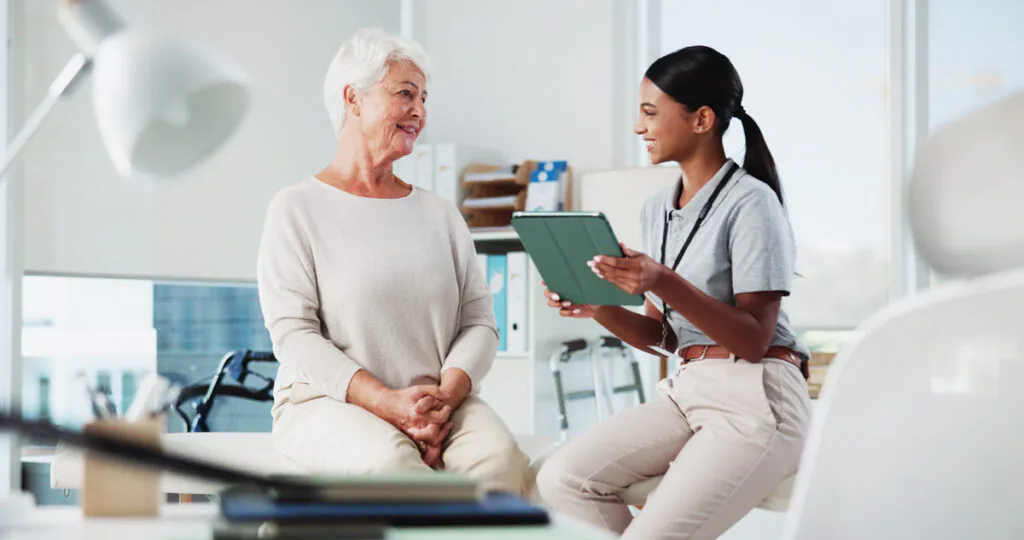 An older woman and a younger healthcare professional with a tablet, smiling and talking in a clinic.
