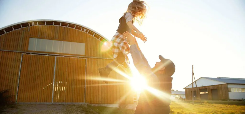Caucasian young handsome father playing with small cute daughter and tossing up in air. Outdoor in sunlight. Farming lifestyle. Man having fun and throwing up kid at farm. Summer at countryside.