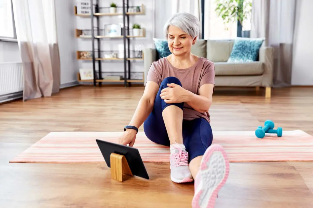 An older woman sits on a yoga mat, looking at a tablet, with dumbbells nearby, in a home setting.