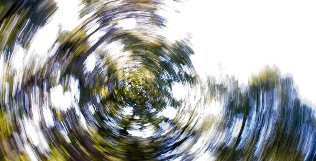 Abstract image with a strong circular motion blur, showing swirling streaks of green, blue, and white, resembling blurred trees and sky.