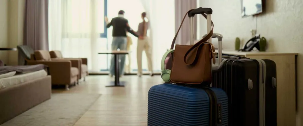 Wide angle view of young couple arriving in hotel room, unpacking luggage and starting vacation stay, checking view from window. Travel booking, hospitality marketing, family tourism promotion