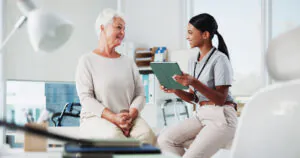 Older woman smiling with physical therapist reviewing recovery plan on tablet" (the site's current alt text — "An older woman with white hair smiles at a younger woman holding a green tablet, both in a bright office setting as she explains Pain Neuroscience Education