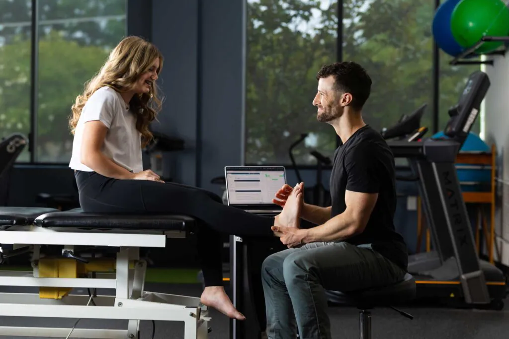 A male physical therapist examines the foot of a female patient seated on an examination table in a clinic, with a laptop and gym equipment in the background.