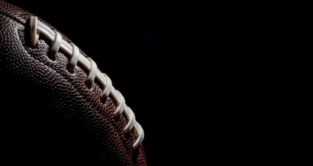 Close-up of a brown American football with white laces against a black background.
