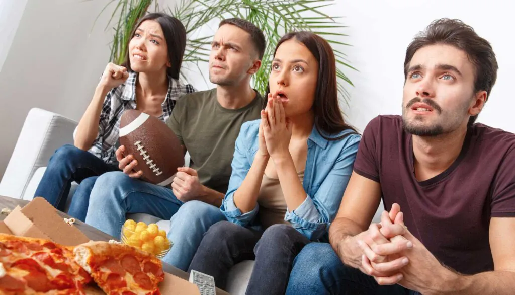 Four young adults on a sofa, looking anxious and focused while watching a game, with a football, pizza, and snacks on a coffee table.