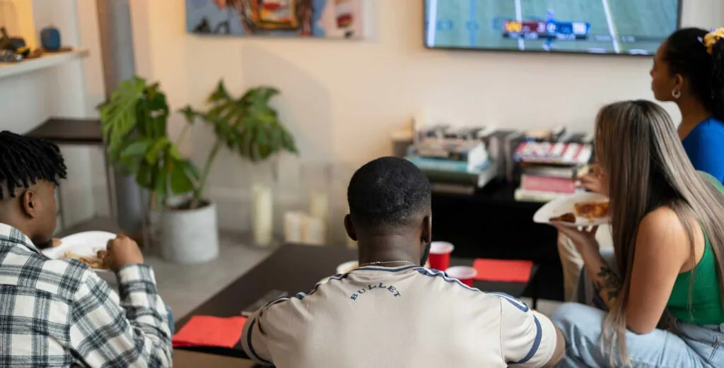 Four people viewed from behind in a living room, watching a sports game on a TV while eating.