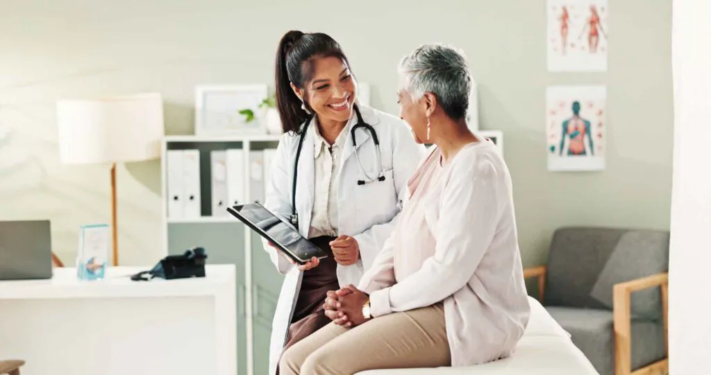 A smiling female doctor in a white coat shows a tablet to an older, smiling female patient seated on an examination table.