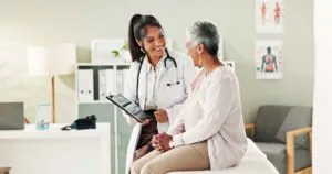 A smiling female doctor in a white coat shows a tablet to an older, smiling female patient seated on an examination table.