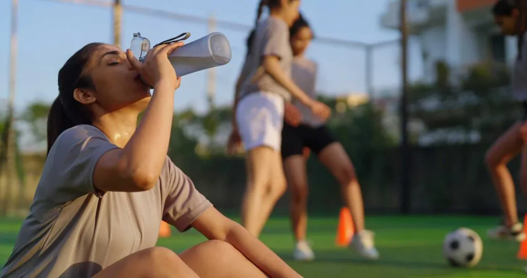 A young woman, sweating, drinks from a water bottle on a green sports field with other athletes in the blurred background.