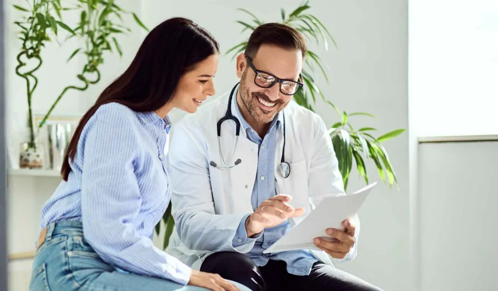 A smiling male doctor in a white coat and a female patient in a striped shirt look at documents together.