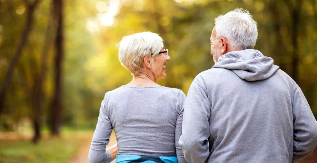 Rear view of an older man and woman walking outdoors, the woman looking back and smiling at the man.