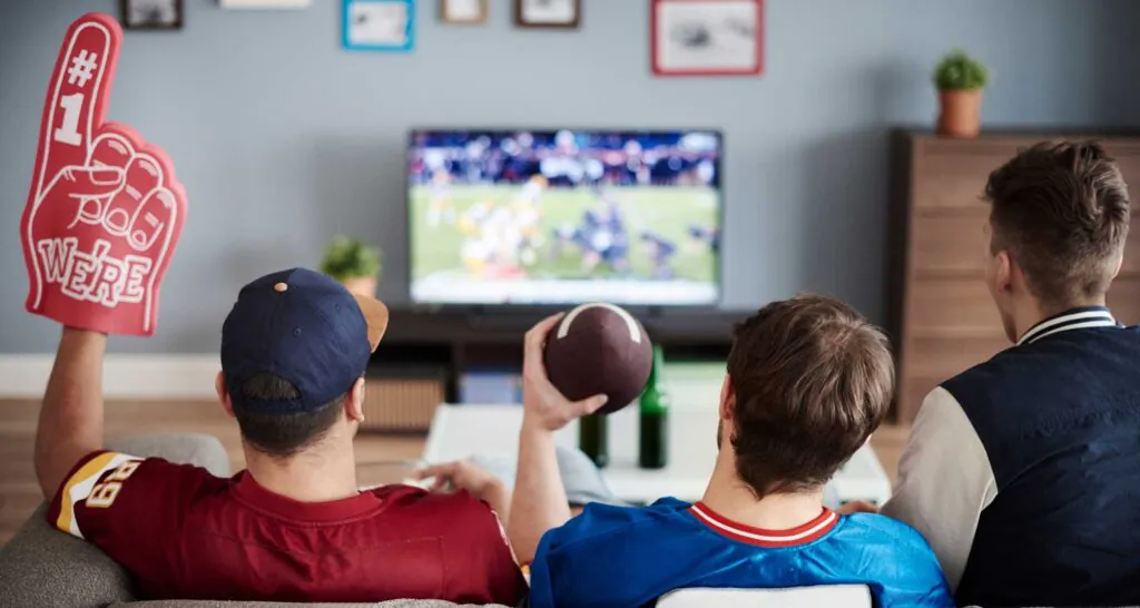 Three people seen from behind, sitting on a couch watching a fantasy football game on TV. One holds a red foam finger, another a small football.