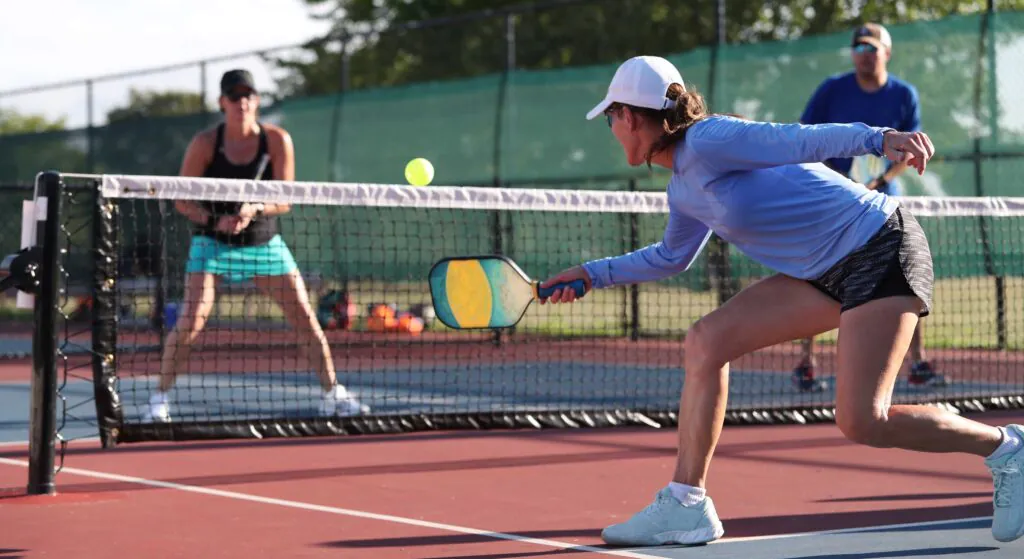 A woman in a white cap and blue shirt lunges to hit a yellow pickleball over the net during a game, with other players in the background.
