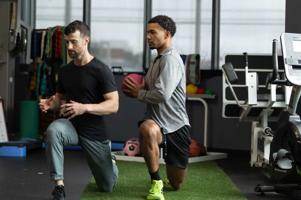 Two men performing lunges in a gym, one holding a pink medicine ball.