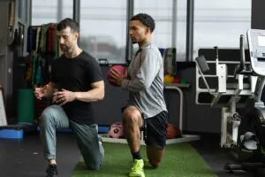 Two men performing lunges in a gym, one holding a pink medicine ball.