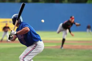 A baseball batter in a blue jersey and helmet holds a bat, facing a blurred pitcher who has just thrown the ball, which is visible in mid-air.