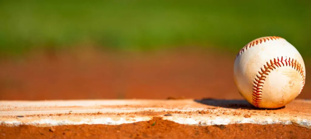 A baseball with red stitching sits on a white chalk line on a dirt baseball field. The background is blurred green grass.