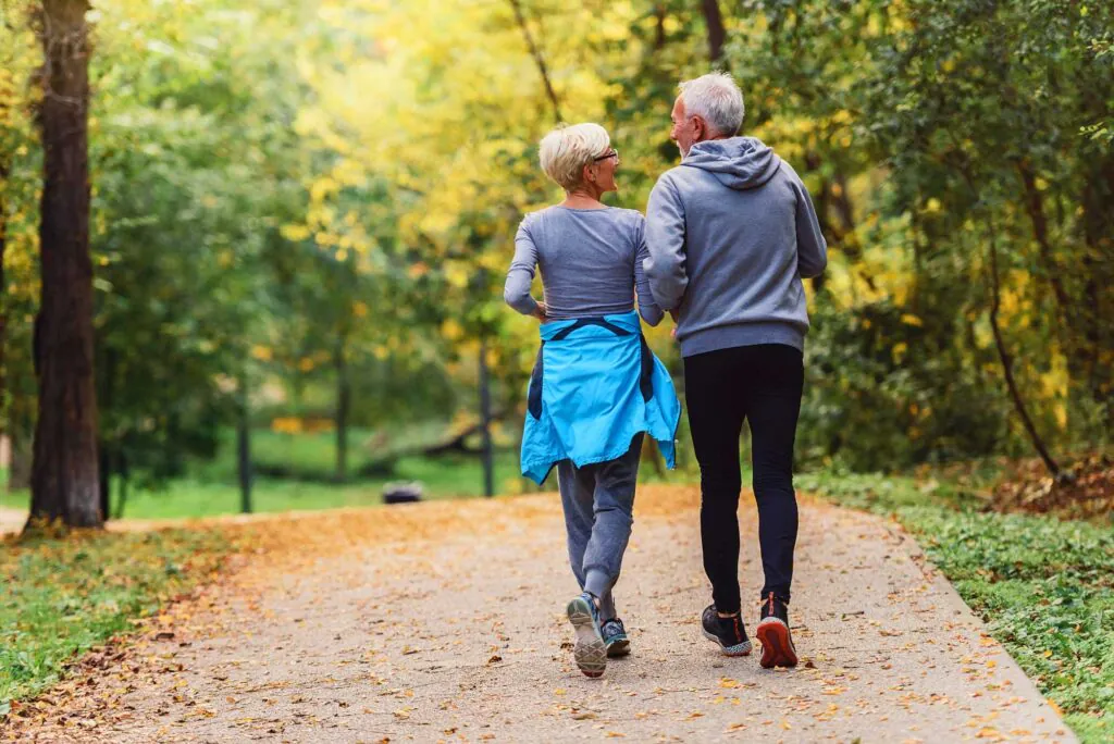 Rear view of an older couple walking on a leaf-strewn path in an autumn park. The woman wears a blue jacket around her waist, and the man wears a grey hoodie.