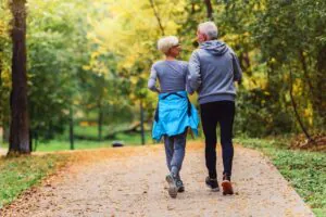 Rear view of an older couple walking on a leaf-strewn path in an autumn park. The woman wears a blue jacket around her waist, and the man wears a grey hoodie.