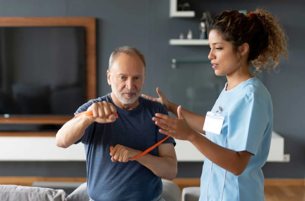 An older man exercising with an orange resistance band, guided by a female physical therapist in a light blue uniform.