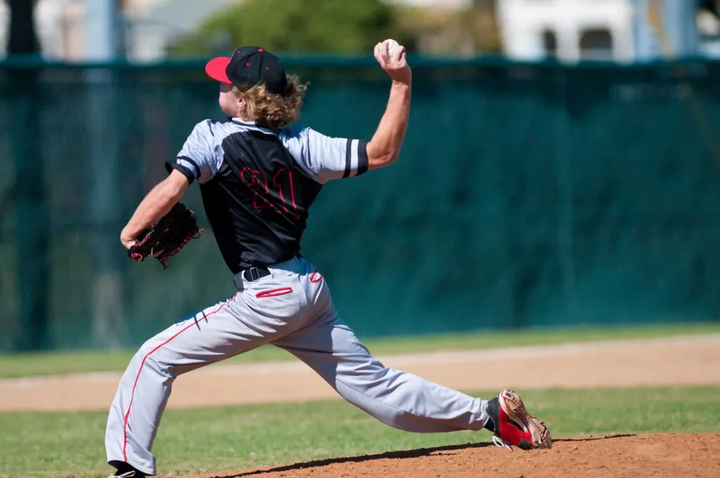 A baseball pitcher in a black and gray uniform with red accents, number 21, throws a ball from a dirt mound on a sunny field.