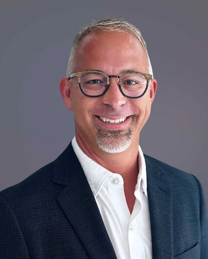 Headshot of a smiling man with gray hair, goatee, and glasses, wearing a dark blazer and white shirt.