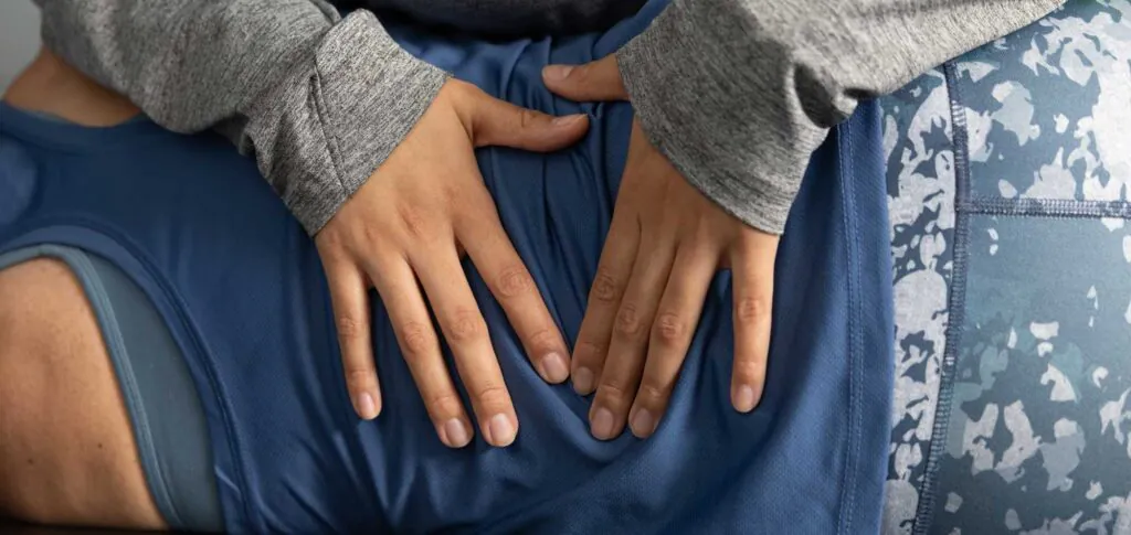 Close-up of two hands in a grey long-sleeved shirt gently touching a person's lower back or hip area, who is wearing blue athletic shorts.