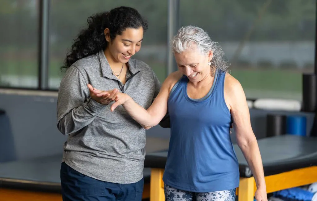 A physical therapist in a gray shirt gently guides an older woman in a blue tank top, both smiling.