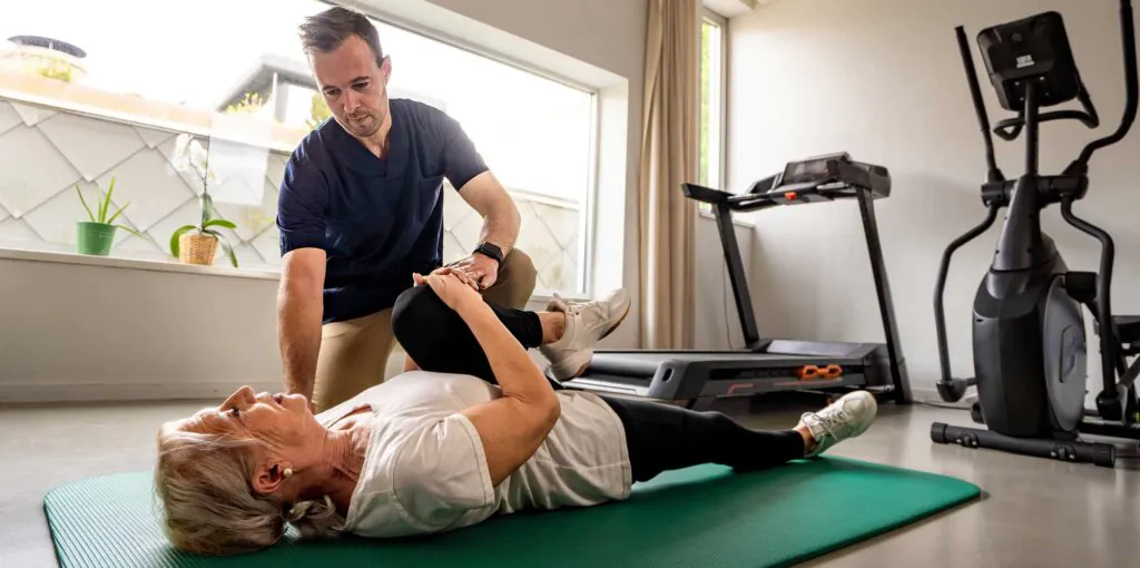 A male physical therapist helps an older woman stretch her leg while she lies on a green mat, in a bright room with exercise equipment.